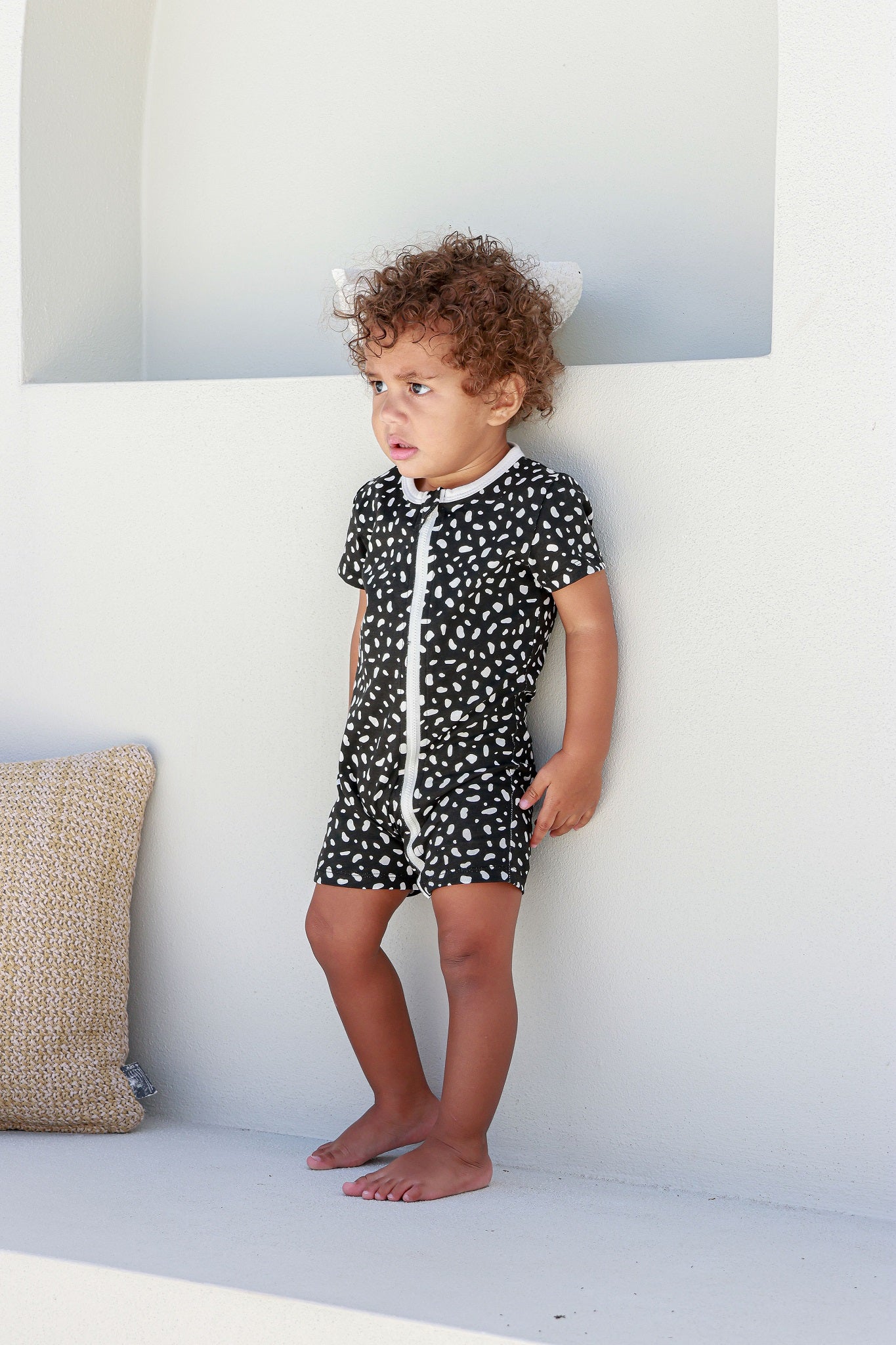 boy toddler standing against white wall wearing black and white spotted romper