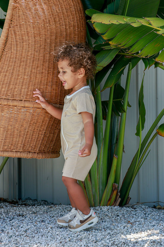 Toddler boy wearing logo beige romper standing near swinging egg chair