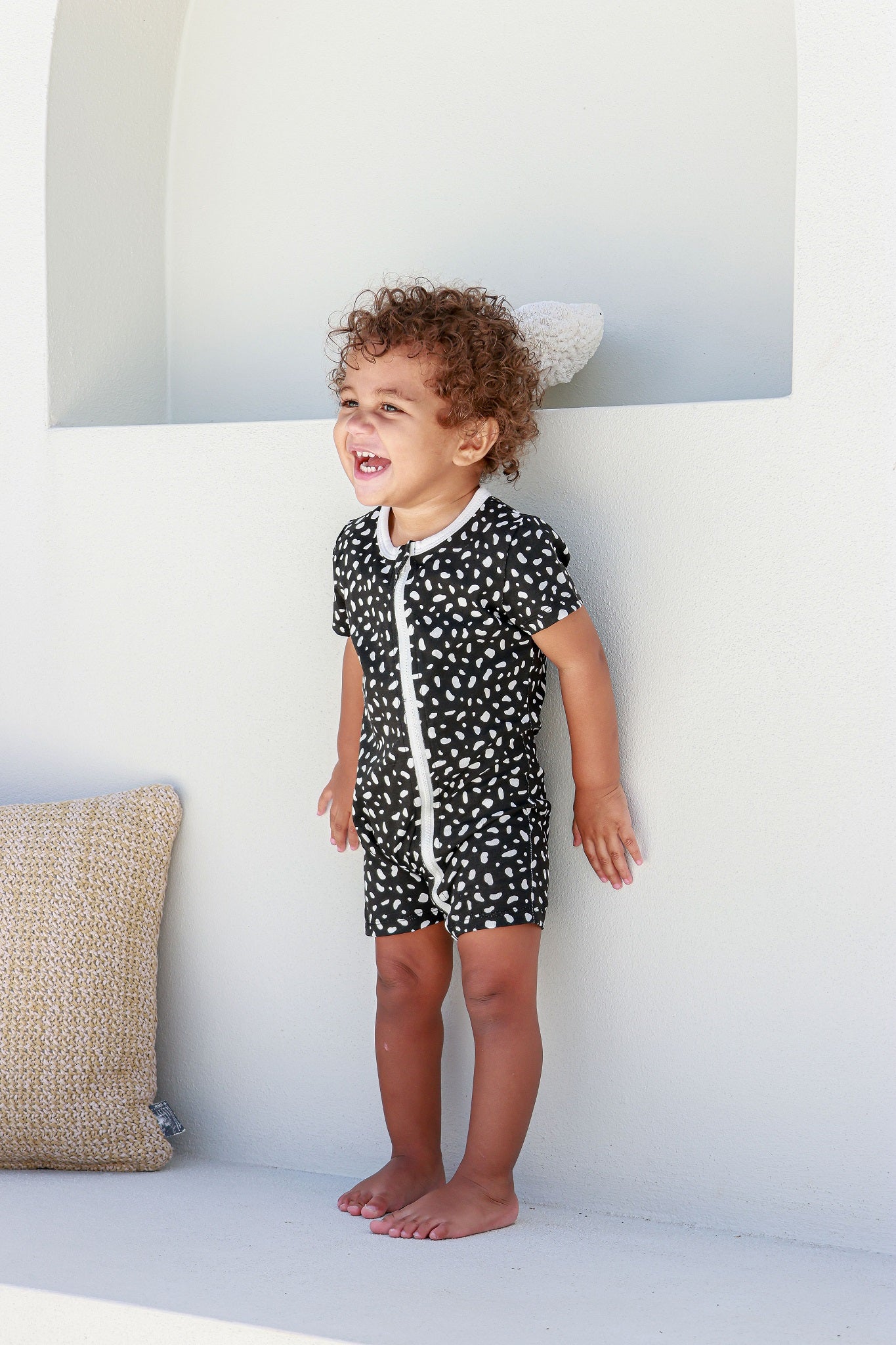 boy toddler standing against white wall wearing black and white spotted romper