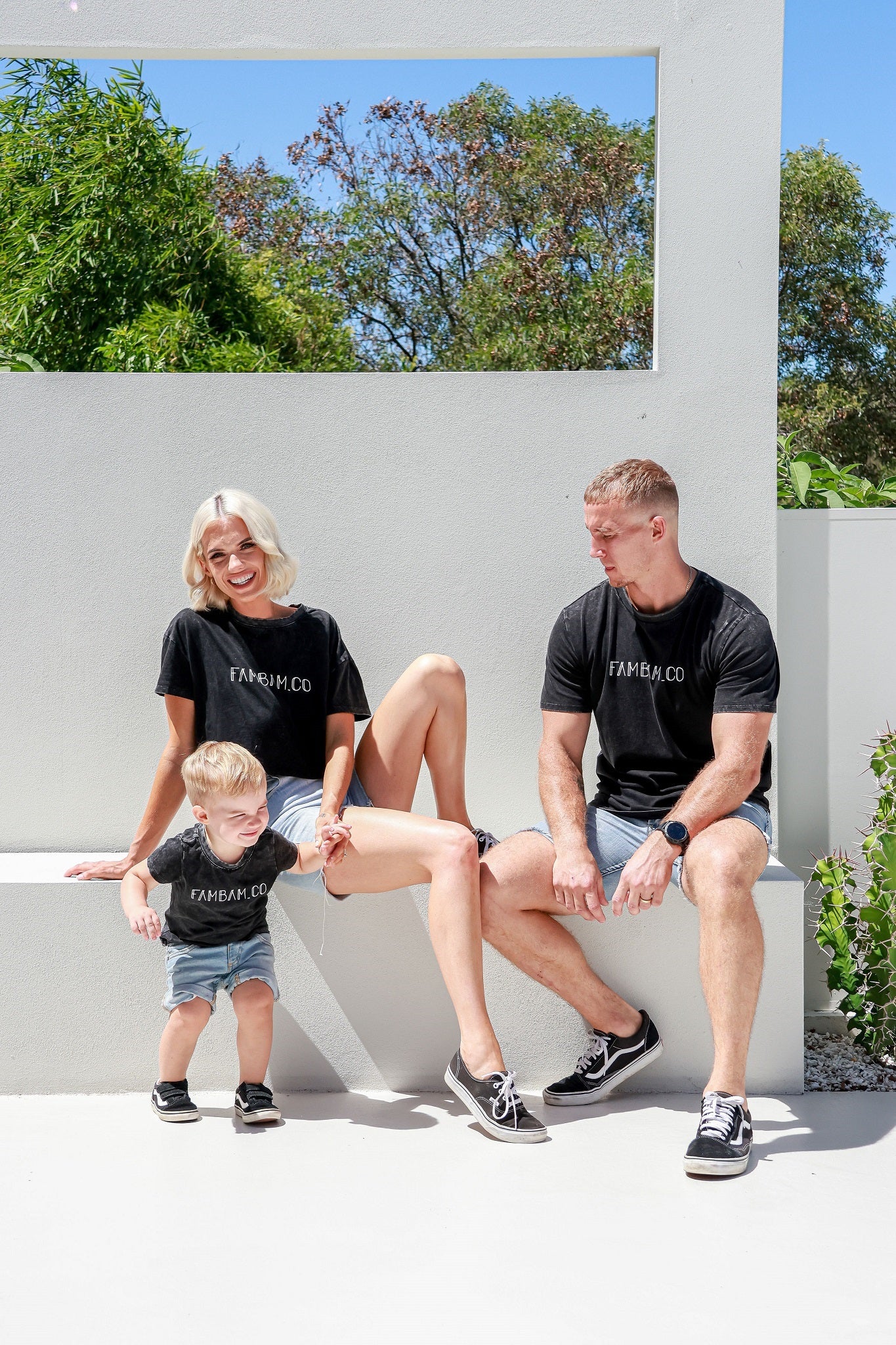 Family laughing and smiling sitting down on white seat holding toddler boys hand while wearing matching acid wash shirts