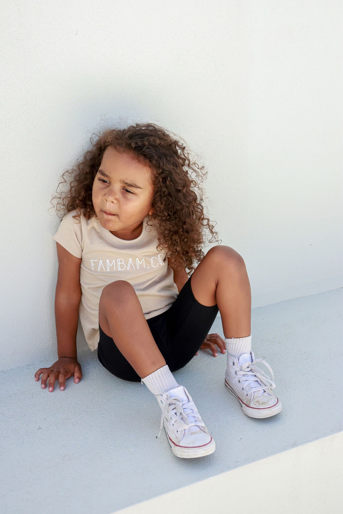 toddler girl sitting posing in front of white backdrop wearing beige logo shirt