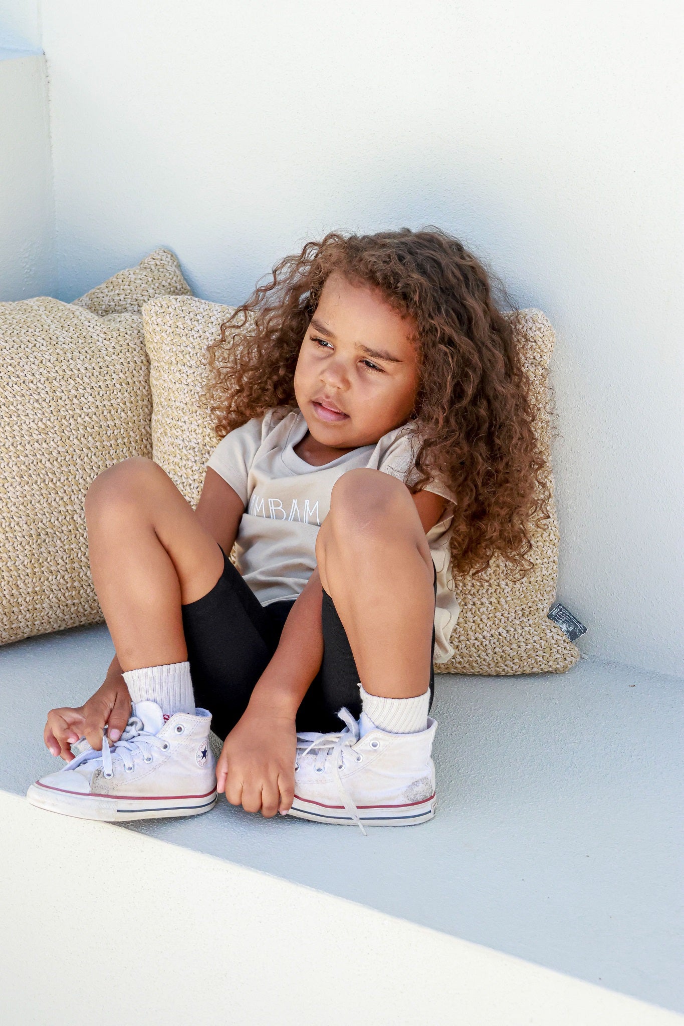 toddler girl sitting posing in front of white backdrop wearing beige logo shirt