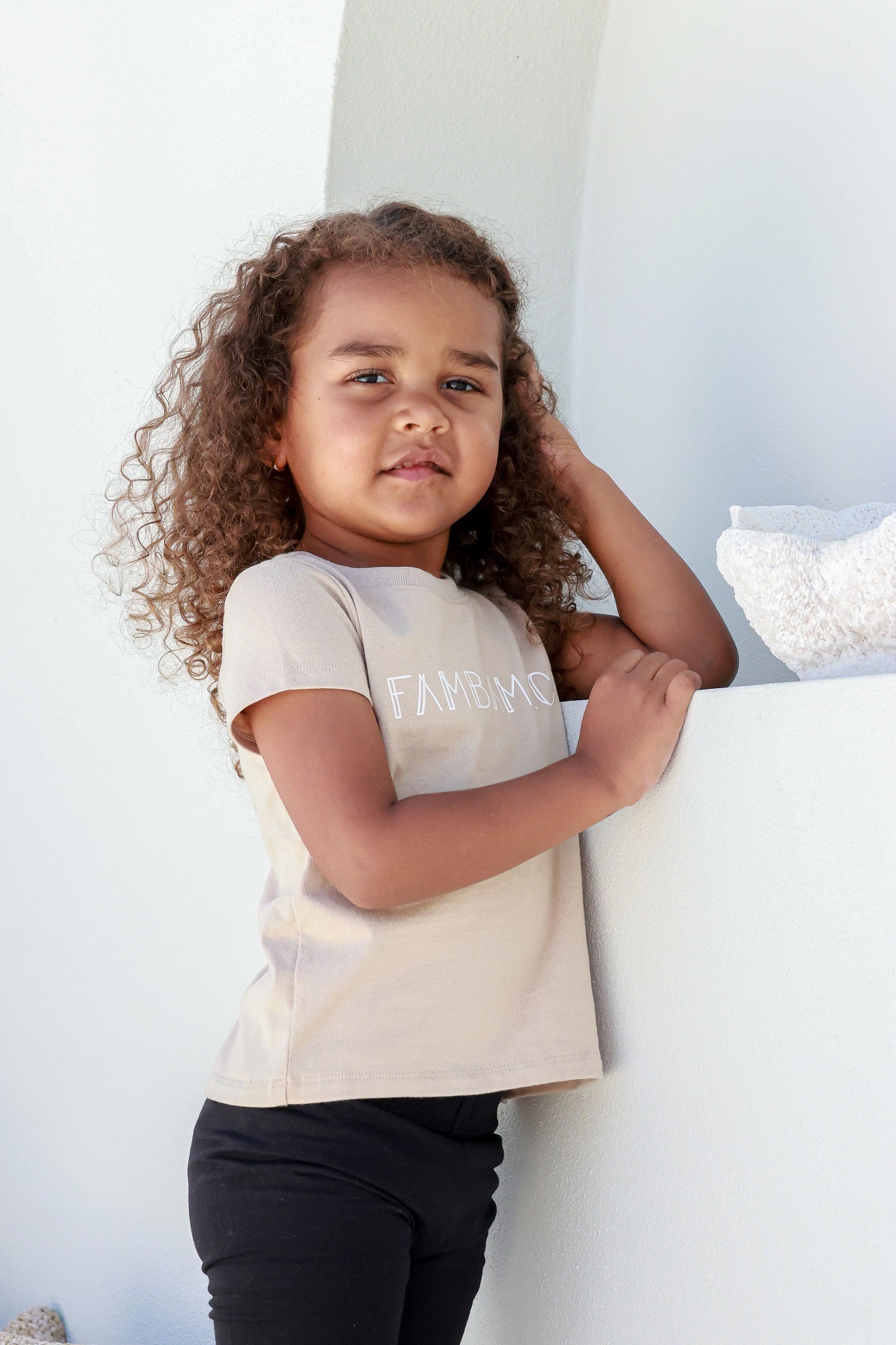 toddler girl standing posing in front of white backdrop wearing beige logo shirt