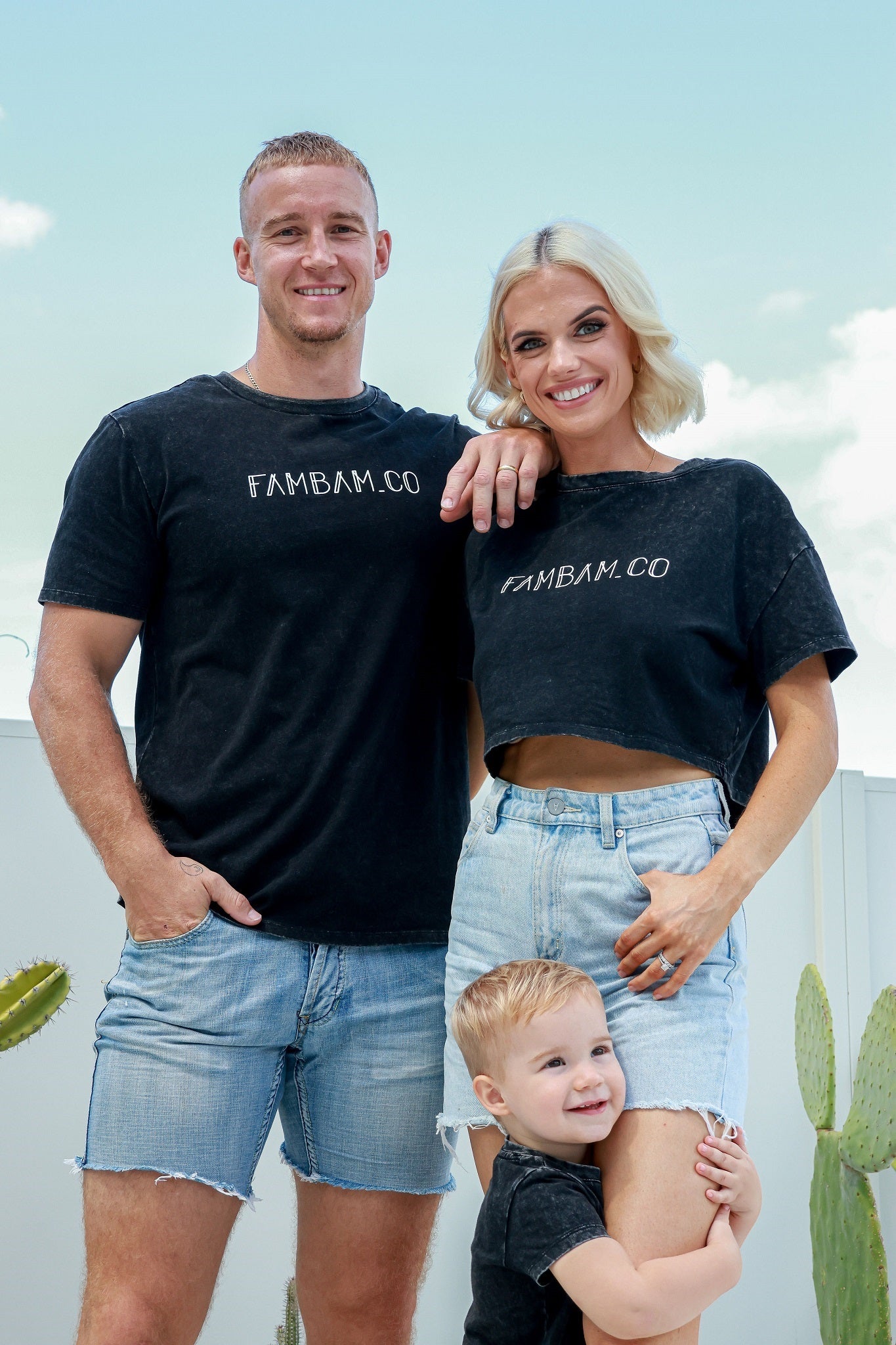 Family standing together smiling toddler hugging mums leg while wearing matching acid wash shirts