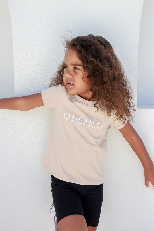 toddler girl standing posing in front of white backdrop with a foot up on wall wearing beige logo shirt