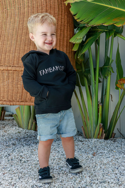 Toddler boy standing outside and smiling wearing black logo jumper 