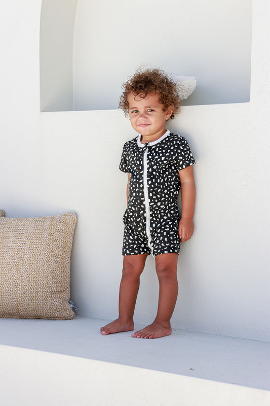 boy toddler standing against white wall wearing black and white spotted romper